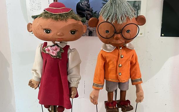 Two puppets, one with a red dress and hat and the other with an orange shirt, sit on a display in front of a white wall.