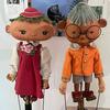 Two puppets, one with a red dress and hat and the other with an orange shirt, sit on a display in front of a white wall.