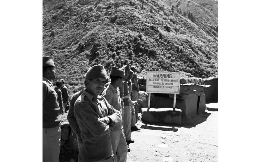 A black and white photo of security personnel standing in a line at a checkpoint, with hills in the distance.