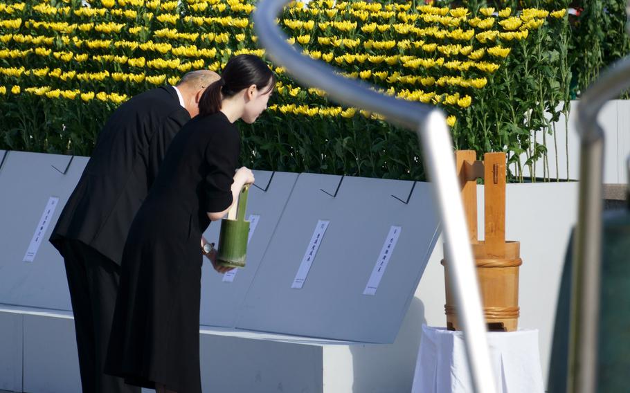 A man and woman, holding a bamboo bucket of water, bow in front of a memorial.