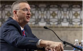 Collins is seated and speaking into a microphone as he points his index finger of his right hand into the table.