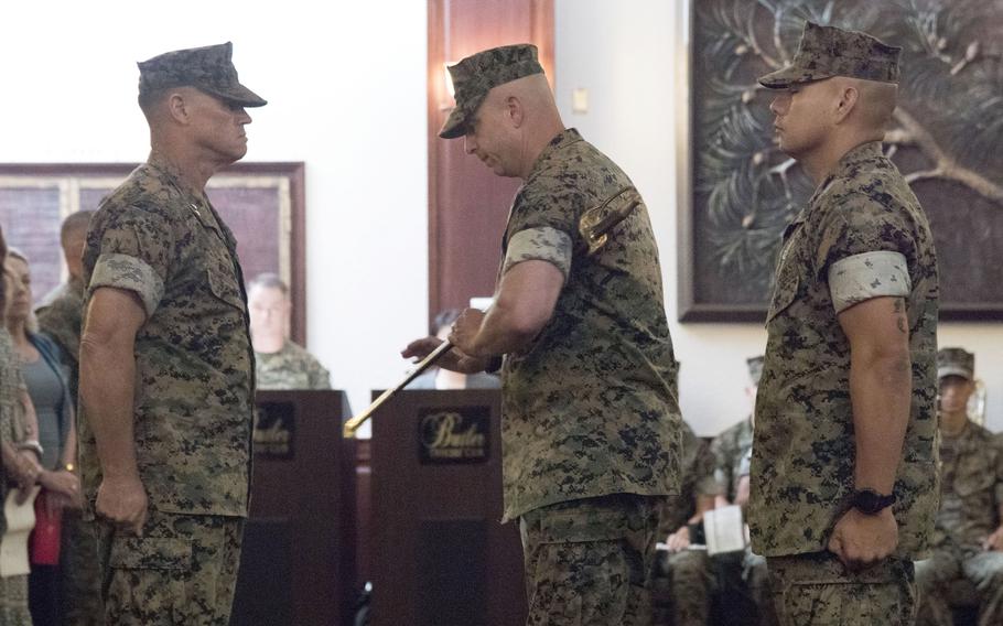 Two Marines in camouflage uniforms and hats stand at attention as a third Marine in camouflage uniform and hat stands between them to hand off a ceremonial sword.