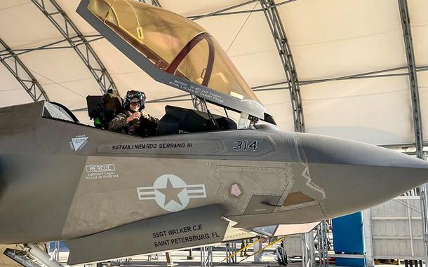 An aviator in flight gear sits in the cockpit of a military fighter jet in a hangar.