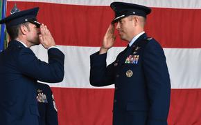 Two Air Force officers in blue dress uniforms face each and salute in front of a large U.S. flag during a change-of-command ceremony.