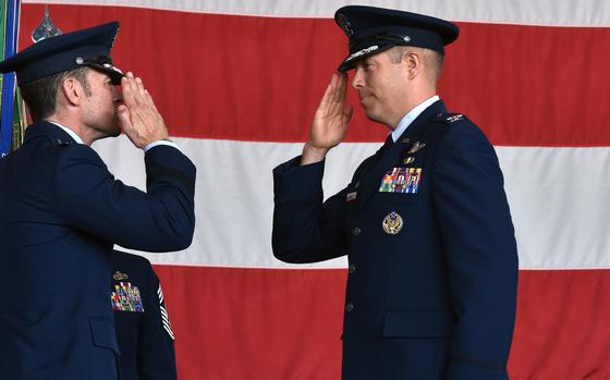 Two Air Force officers in blue dress uniforms face each and salute in front of a large U.S. flag during a change-of-command ceremony.