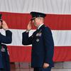 Two Air Force officers in blue dress uniforms face each and salute in front of a large U.S. flag during a change-of-command ceremony.