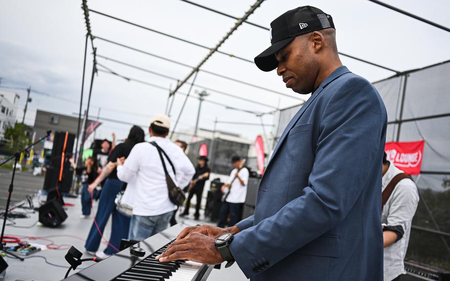 A man in a blazer and baseball hat, seen from the side, plays a keyboard on a stage with other musicians behind him.