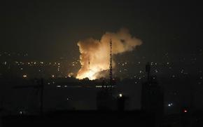 An illuminated plume of smoke rises into the sky over a darkened city skyline at night.