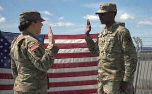 An Army soldier raises their right hand while taking an oath in front of another soldier administering the oath. Both are dressed in camouflage uniforms and hats, standing outdoors with a large U.S. flag displayed behind them.