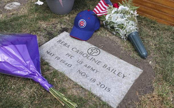 A stone grave marker reading “Reba Caroline Bailey, PFC U.S. Army, May 19 1940, Nov. 18 2015” is seen from above, with two bouquets of flowers and a blue Chicago Cubs baseball hat on the ground next to the marker.