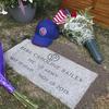 A stone grave marker reading “Reba Caroline Bailey, PFC U.S. Army, May 19 1940, Nov. 18 2015” is seen from above, with two bouquets of flowers and a blue Chicago Cubs baseball hat on the ground next to the marker.