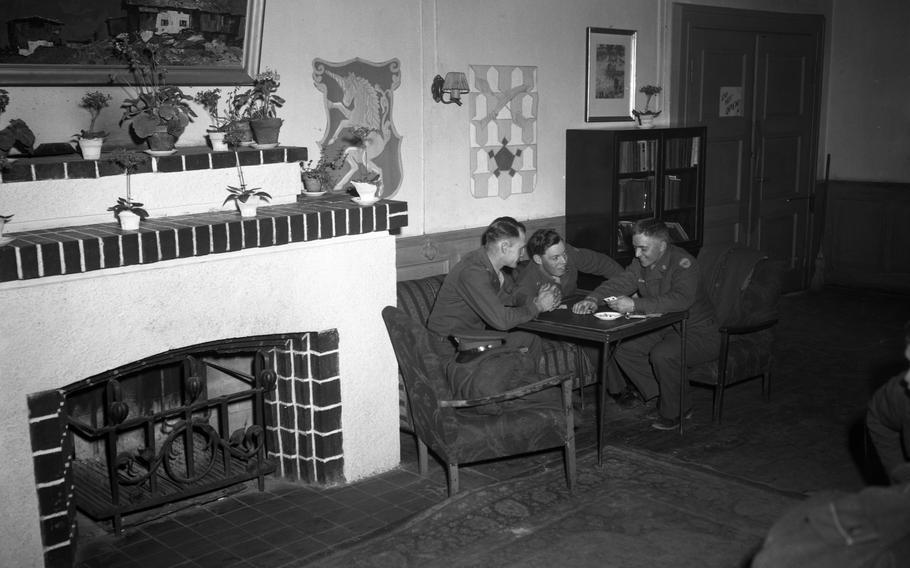 A room with a fireplace, in which three smiling soldiers lean around a small table.