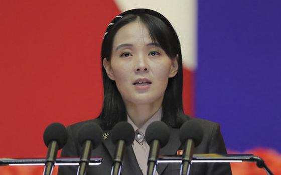 A Korean woman in a dark suit speaks into multiple microphones while standing at a podium, with red, white and blue colors visible in the blurred background.