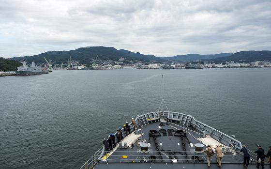 A view from the bow of a naval vessel showing the forward deck with crew members positioned along the railings. The ship is entering or leaving a harbor surrounded by forested mountains.