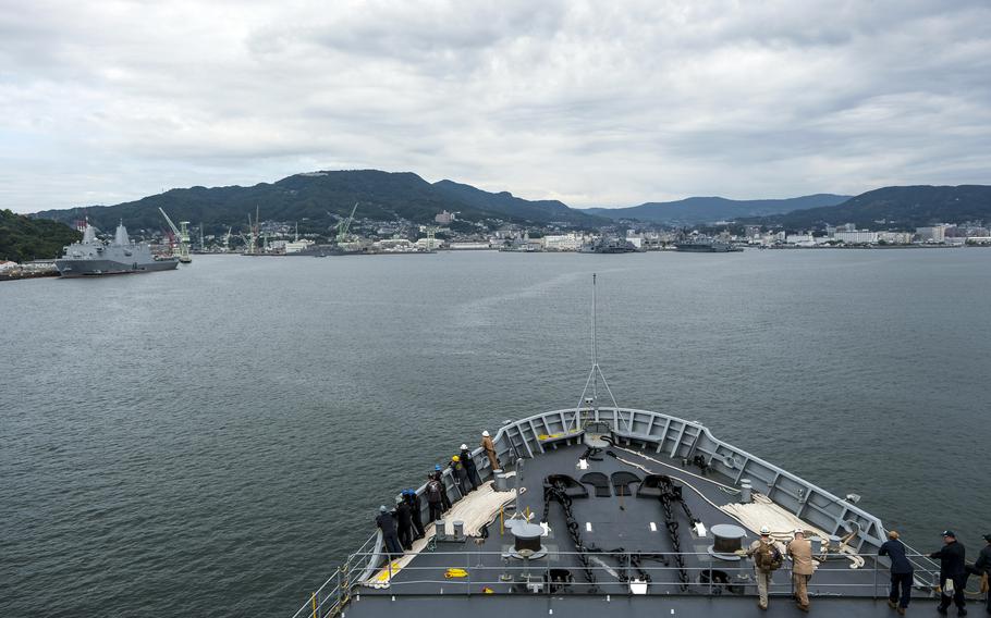 A view from the bow of a naval vessel showing the forward deck with crew members positioned along the railings. The ship is entering or leaving a harbor surrounded by forested mountains.