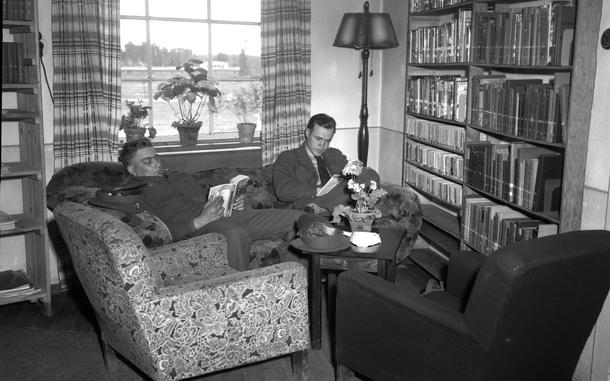 Two men recline on a couch and read books in the corner of a library room.