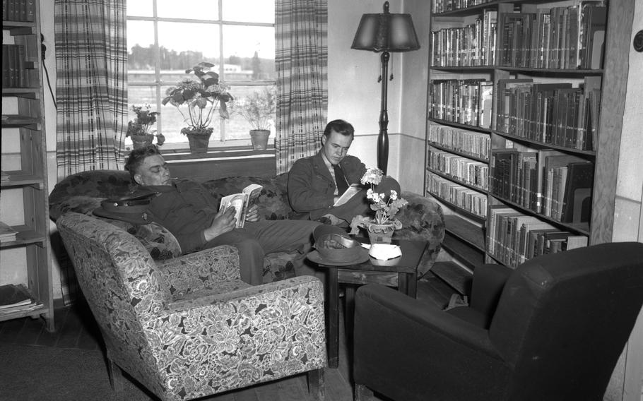 Two men recline on a couch and read books in the corner of a library room.