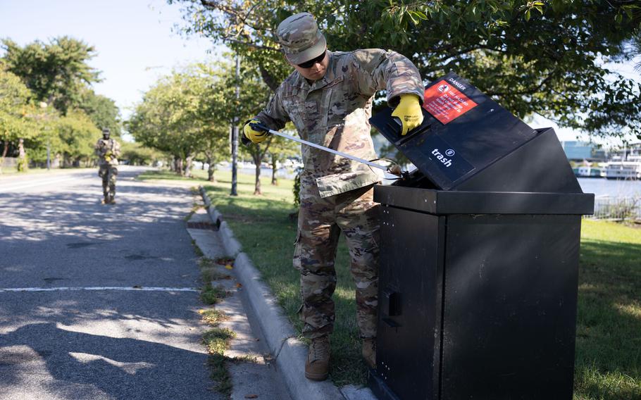 A soldier with gloves opens a trash bin to drop in something he collected up with his trash picker.