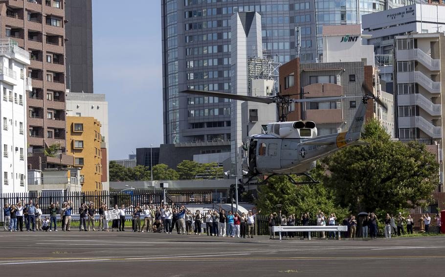 A large crowd of people line up along a helicopter landing area, many holding signs and looking up at the sky. A U.S. military helicopter is positioned on the right side of the frame. 