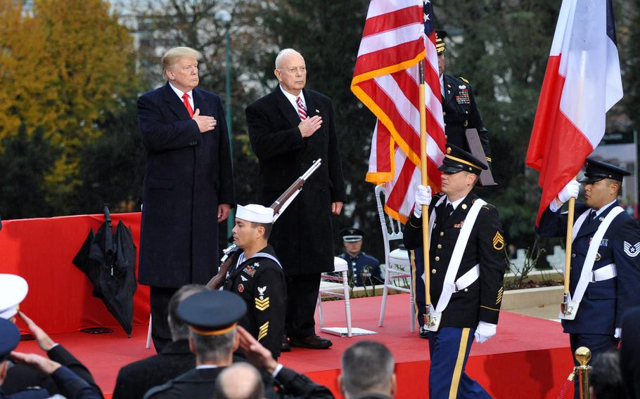 President Donald Trump stands wiht his hand over his heart in front of an American flag as a color guard passes by in front of him.