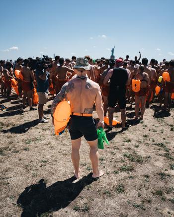 A man stands with his back to the camera holding an orange flotation device. In the background are hundred of other NYC SEAL Swim participants in matching black swim trunks. The Statue of Liberty can be seen in the distance.
