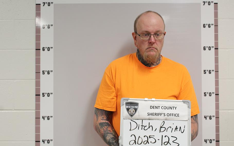 A man wearing an orange shirt stands to have his mugshot taken in jail.