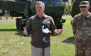 A man in an olive-green button-down shirt speaks into a microphone while standing in a grassy area next to an Army officer in camouflage uniform and in front of an armored vehicle.