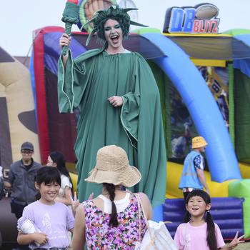 A woman on stilts and dressed as the Statue of Liberty poses with two children standing on either side of her as another woman, seen from behind, stands in front of them to take a photo at an outdoor festival.