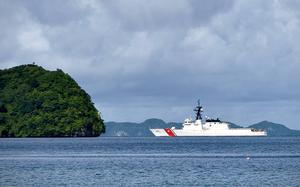 A Coast Guard ship, seen from a distance, sails in open water with a forested outcropping of land to its left and the horizon in the background.