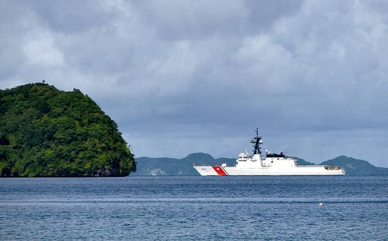 A Coast Guard ship, seen from a distance, sails in open water with a forested outcropping of land to its left and the horizon in the background.