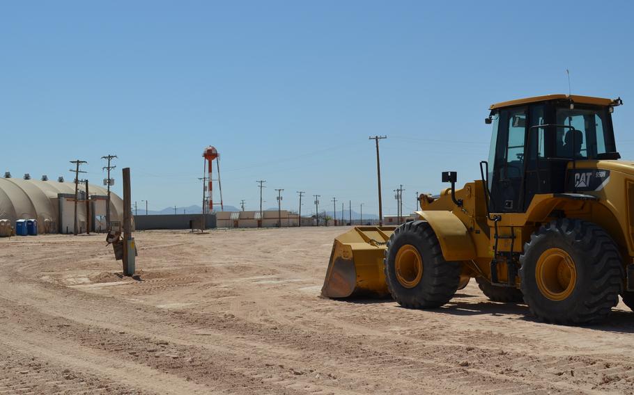 A bulldozer sits on an empty site with some structures in the background.