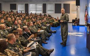 A military officer in flight suit stands presenting to a large seated audience of uniformed service members in an auditorium. 