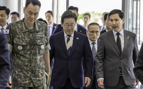 A group of South Korean officials walking together in a corridor of a government building. In the foreground, a military officer in camouflage uniform walks alongside several men in dark business suits. Behind them, additional officials and staff members follow.