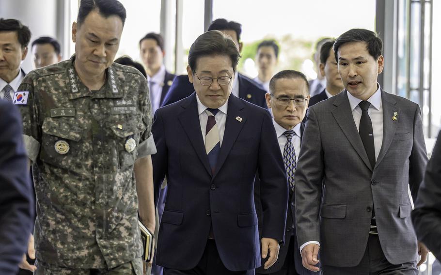 A group of South Korean officials walking together in a corridor of a government building. In the foreground, a military officer in camouflage uniform walks alongside several men in dark business suits. Behind them, additional officials and staff members follow.
