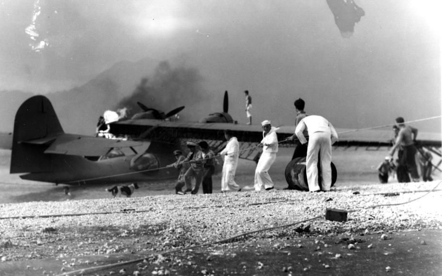 A black-and-white, 1940s-era image of soldiers and sailors in uniform using a rope to pull a damaged airplane from water as smoke rises from the plane.