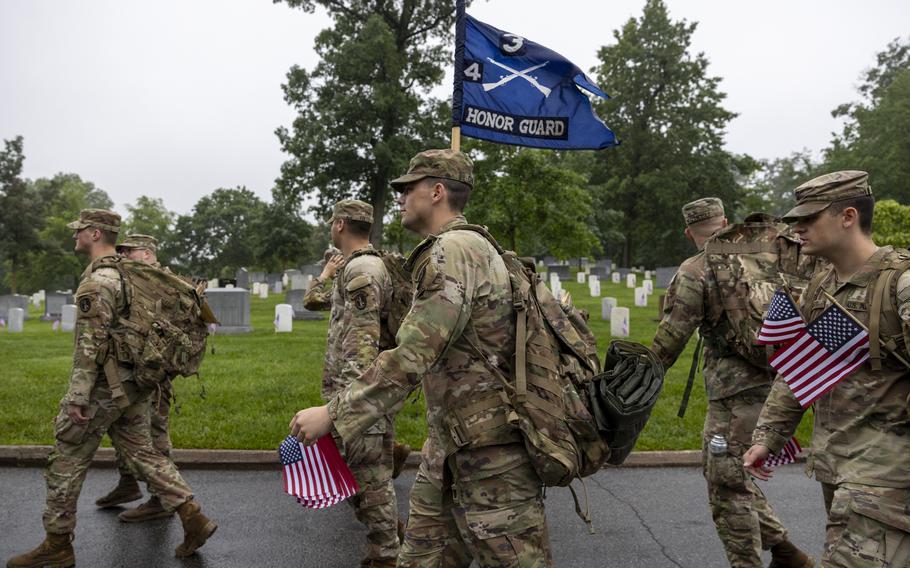 A group of soldiers carry American flags as they walk down the road before placing the flags at headstones. One soldier also carries a flag that says Honor Guard.