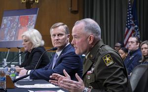 A man in an Army uniform gestures as he speaks into a microphone on a table.