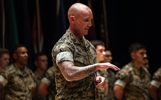 A U.S. Marine in camouflage uniform stands in the foreground, gesturing with his right hand while speaking. He has a shaved head and visible sleeve tattoo on his right arm. Behind him, a formation of fellow Marines stands in rows, also wearing camouflage uniforms.  The lighting highlights the speaker while the others remain slightly out of focus.