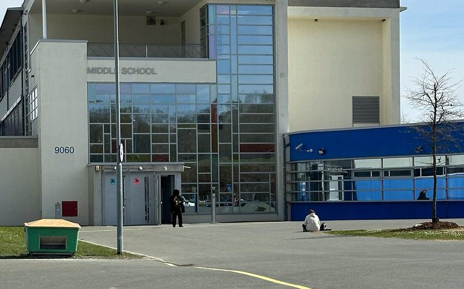 The distant figures of two people are seen outside of a school building, one sitting on the ground.