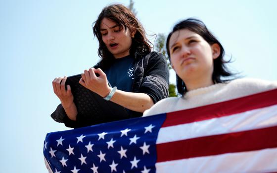A teenaged girl reads from a tablet, while another stands in front of her holding a U.S. flag.