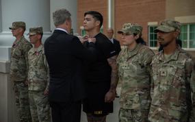 A man in a suit seen from behind, pinning a medal onto a soldier who stands at attention.