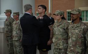 A man in a suit seen from behind, pinning a medal onto a soldier who stands at attention.