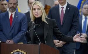 Pam Bondi stands at a podium and speaks into a microphone as other government officials stand behind her.