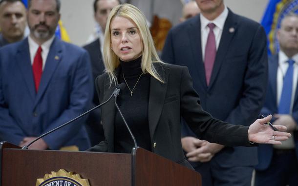 Pam Bondi stands at a podium and speaks into a microphone as other government officials stand behind her.
