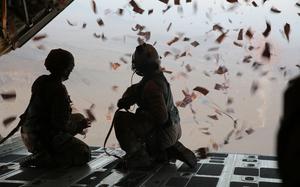 Two service members in flight suits, seen from behind, kneel on the lowered rear ramp of an aircraft as leaflets fly out the open back of the aircraft and fly into the air.