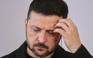 A man with dark hair and a beard holds his hand to his forehead in a gesture suggesting stress, fatigue or deep thought. He’s wearing a dark colored shirt or jacket and looking downward with a contemplative or troubled expression against a neutral background.