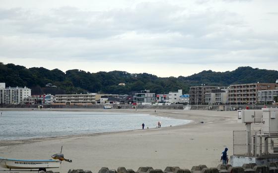 A quiet beachfront with a few people walking dogs along the shore. Low-rise buildings and apartments line the waterfront, backed by green hills under a cloudy sky. A small boat and utility structures are visible in the foreground.