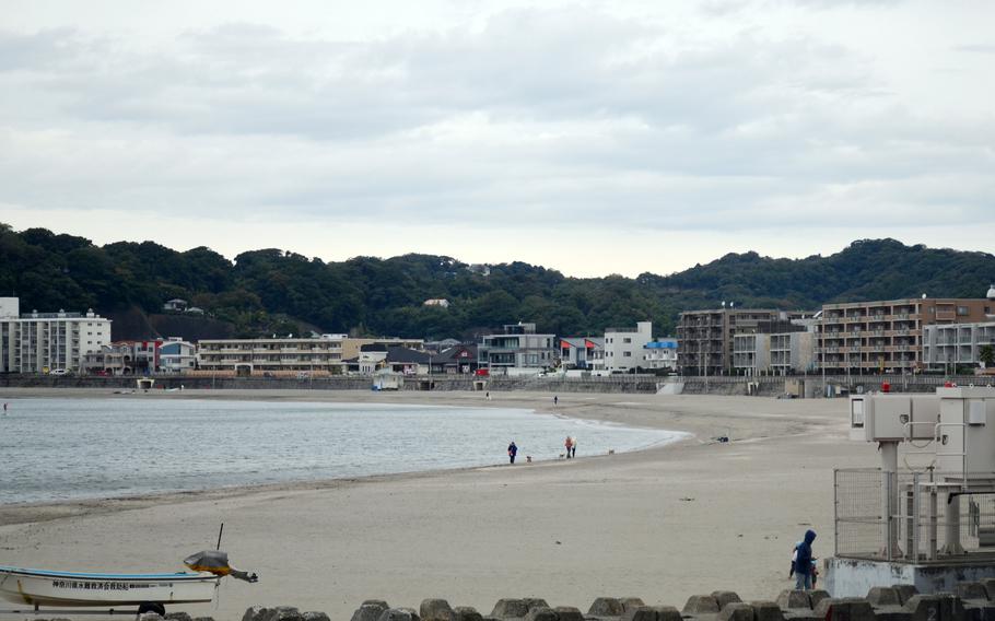 A quiet beachfront with a few people walking dogs along the shore. Low-rise buildings and apartments line the waterfront, backed by green hills under a cloudy sky. A small boat and utility structures are visible in the foreground.