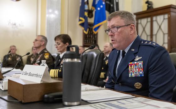 Three military officials in dress uniforms sit at a table in a congressional hearing room with binders and documents in front of them.