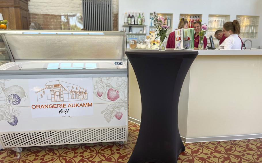 Interior view of a café showing an ice cream display case with “ORANGERIE AUKAMM Café” branding, featuring illustrations of fruits. A tall black cocktail table is positioned nearby, and staff members in red and white uniforms can be seen working in the background.